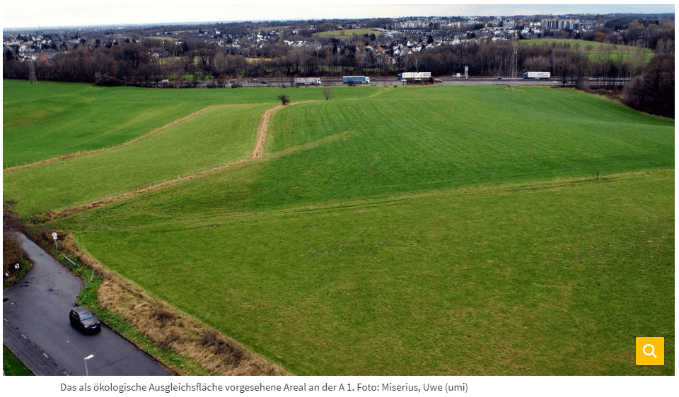 links unten eine Straße mit einem schwarzen parkenden Wagen, dahinter eine große grüne Wiese, dahinterliegendes Wohngebiet wird durch die querlaufenden Autobahn A1 geteilt